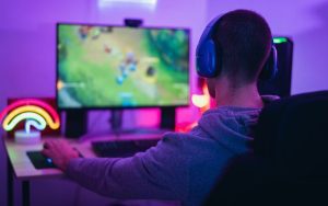 גיימר עם אוזניות משחק במשחק מחשב פופולרי בחדר חשוך מואר באורות ניאון צבעוניים.